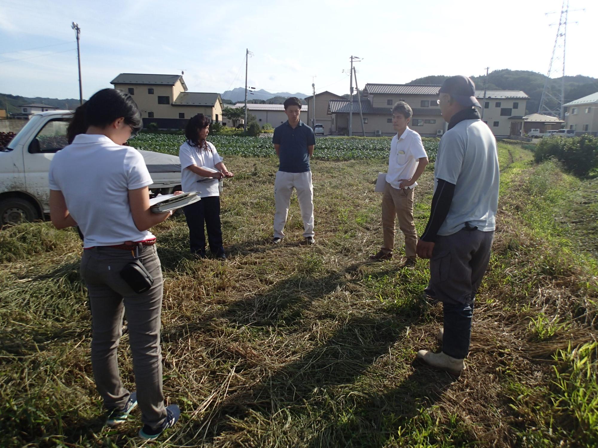 後方に作物が植えられている畑が見える平地で関係者が資料を手にもちながら円になって話をしている写真