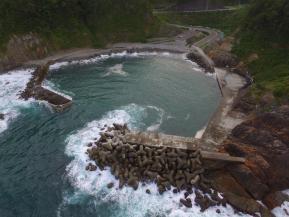 内側に丸くカーブした海岸の両脇から海に突き出す二つの堤防と、堤防の内側の海岸沿いに作られた港を地上に近い上空から見た写真