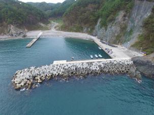 山々を背景に、陸地から海に突き出た堤防に複数の船が停まっている写真