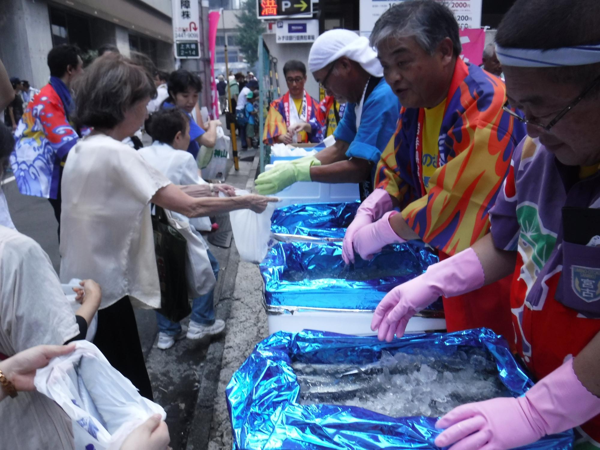 いくつもの箱の中に氷と一緒に入れられたたくさんのさんまを、祭りの来場者に渡している人々の写真