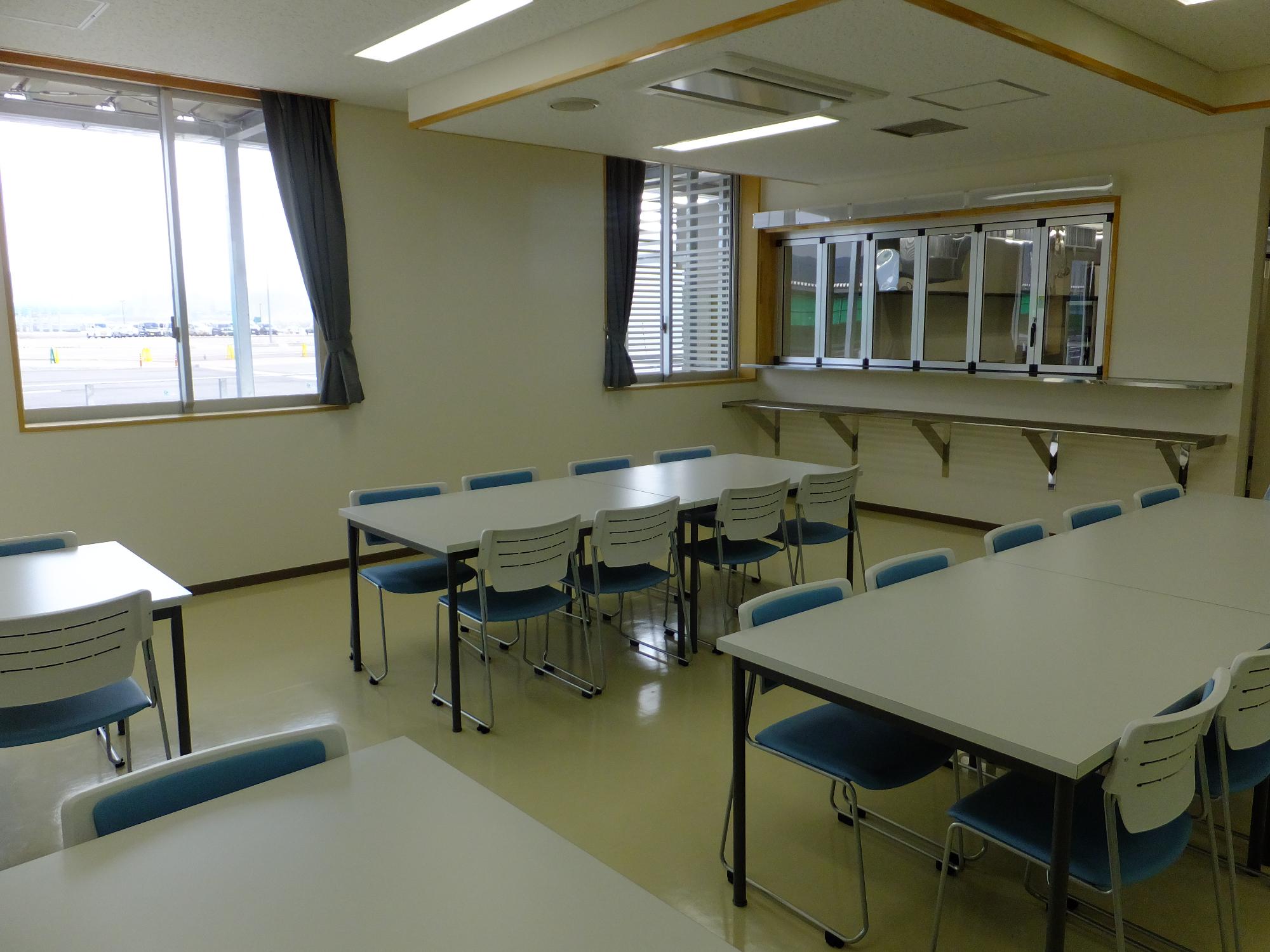 奥に窓ガラス、右に小窓が設けられ室内に8人掛けのテーブルが2列ずつ設置された魚市場食堂内の写真
