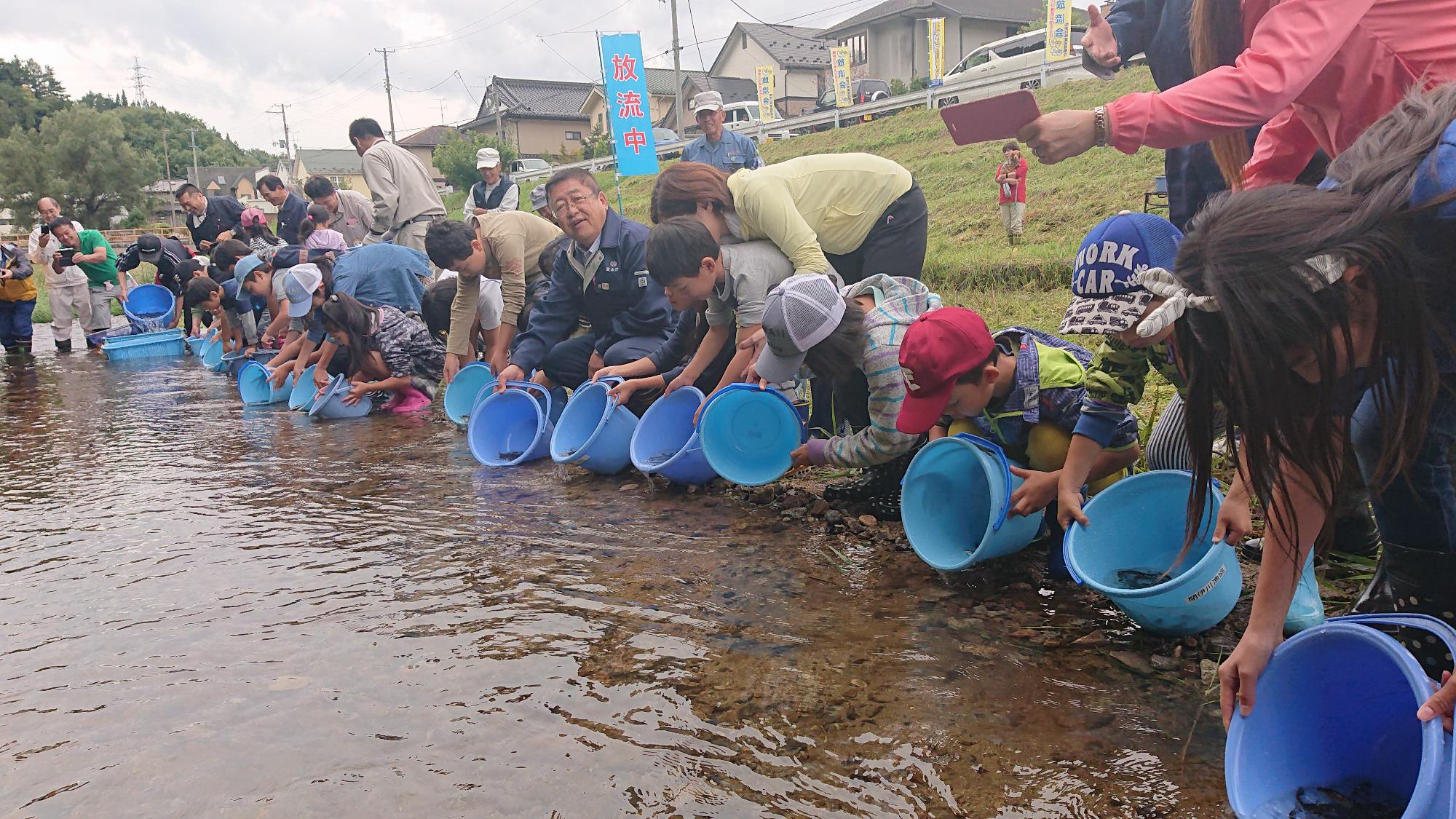 川べりにそって横一列に座ったり前に屈んだりしながら、バケツに入った魚を川に放流している子どもたちや大人たちの写真