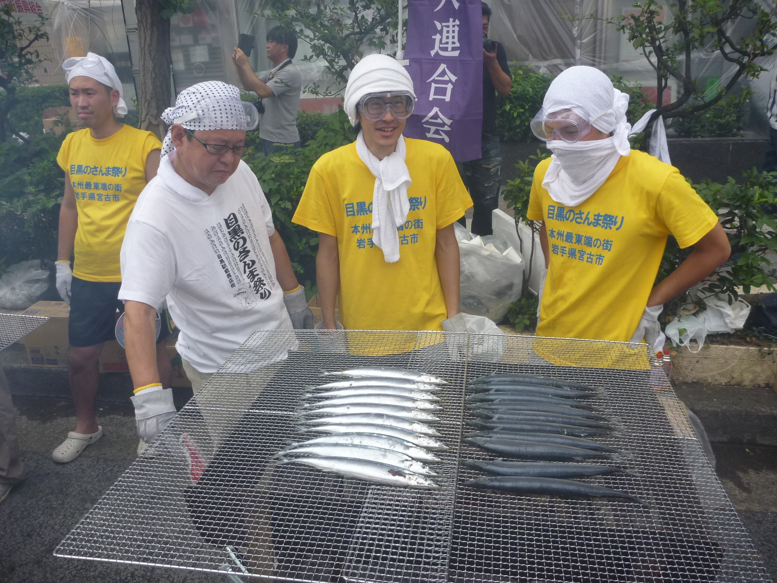 大きな焼き網の上に横二列にたくさんのさんまを並べて焼いている男性三人の写真