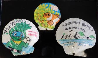 海やお魚のイラストや、日付などの文字が描きこまれたホタテの貝殻が三つ並んでいる写真