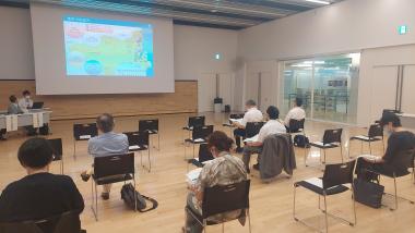 パイプ椅子に参加者の人が座り前方のスクリーンを見ている写真