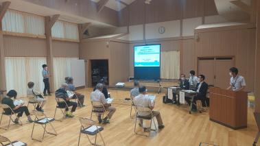 前方のスクリーンを見ながら右側の演台で男性が説明をしている会議の写真