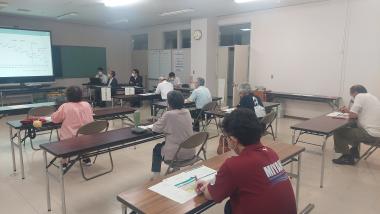 長机が設置されパイプ椅子に座り参加者の人たちがスクリーンを見ている写真