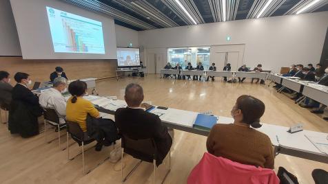 広い室内にコ型に並べた長机に座った関係者の人達が中央のスクリーンを見ている写真