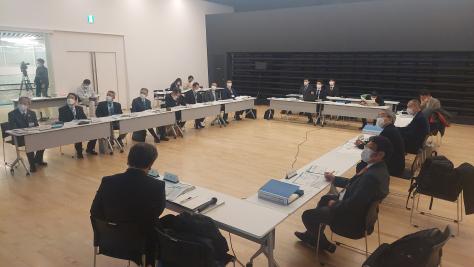 白い長机に会議の参加者の方々が着席している室内の写真