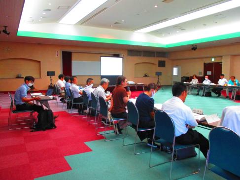 床が赤と水色に分かれている明るい室内で中央の女性がマイクを持ち説明をしている会議の様子の写真