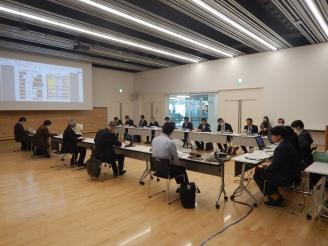 オフホワイトの壁で奥にスクリーンが設置された会議室でコ型に設置された長机に着席している会議参加者の人たちの写真