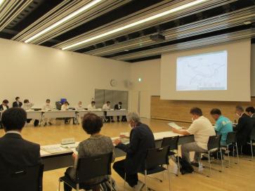 正面のスクリーンに映し出された資料をみながら会議をしている人達を斜め後方から写した写真