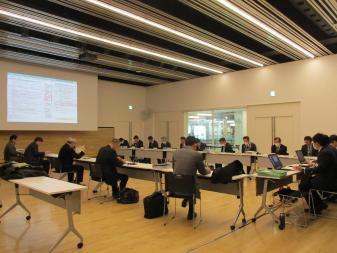 奥のスクリーンに対しコ型に長机を並べ会議出席者が資料をみている室内を写した写真