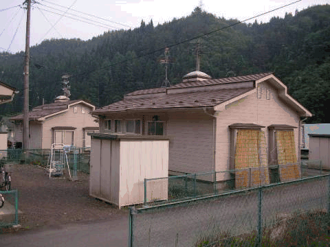 緑に囲まれ、フェンスで仕切られたベージュ色の外壁の飛の沢団地住宅の外観写真