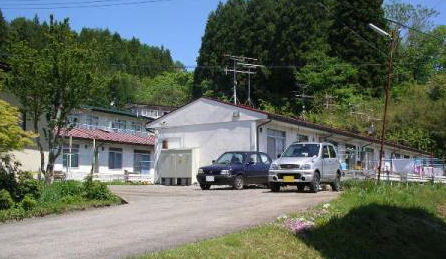 緑に囲まれ、一戸建て住宅が隣り合わせで並んでいる平屋建て津軽石住宅を庭側から撮影した写真