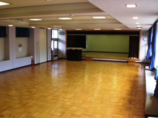 天井に四角い蛍光灯が設置され緞帳のあるステージが前方にある会議室を全体が分かるように撮影された写真