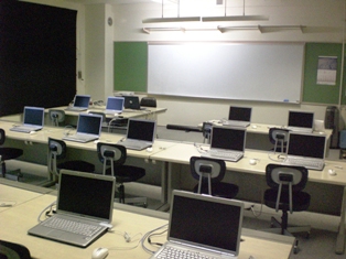 複数を直線で並べた長テーブルが3列ありその上にノートパソコンが数台置かれているコンピューター室の写真