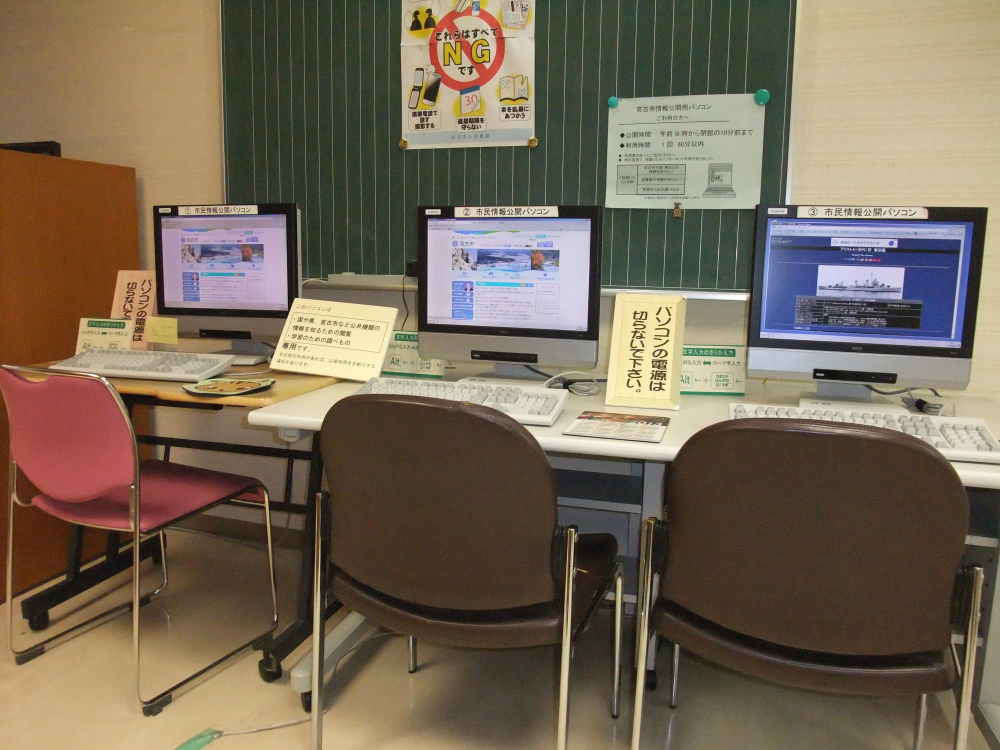 壁に沿ってテーブルが設置されその上にデスクトップパソコンが3台置かれ椅子も3脚設置されている写真
