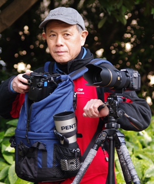 写真家の藤田弘基氏が前に掛けたリュックの中のカメラを右手で取り出しながら、左側にあるカメラの三脚のレバーに手をかけ左方向に目線を向けている写真