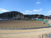 遠くに山々が見え、周りをフェンスで囲まれ照明塔も設置された田老野球場を全体が見えるよう遠目で撮影された写真