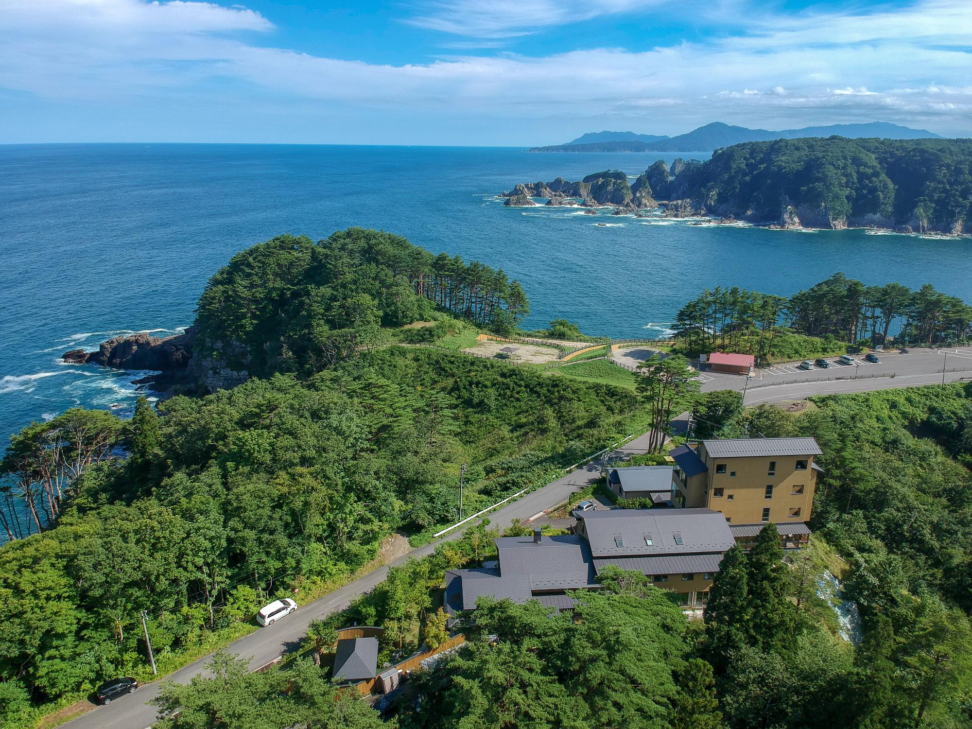 周りを木々に囲まれ道を挟んだ向かい側に広大な海が臨める渚亭たろう庵の建物を上空から撮影した写真