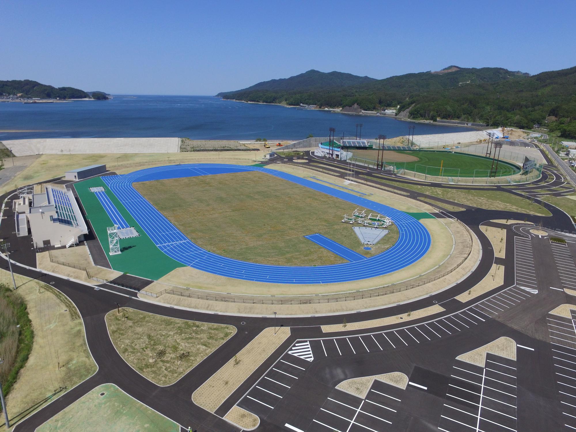 遠くに海と山々が見渡せ、手前に駐車場、中央に芝生が見える宮古運動公園を上空から撮影した写真