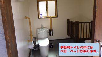 黄色い手すりが水洗トイレ両脇に設置され、トイレ内の一角にベビーベッドが設置された多目的トイレ内の写真