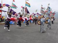 大漁旗がいくつもあがった会場で兜のお面をかぶり旗をひろげている演者と着物を着用し編み笠をかぶり太鼓をかついだ演者が対面になって踊っている様子の写真