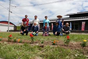 花壇に植えられた赤や黄色の花々の後方に9人が2列に並び記念撮影をしている写真
