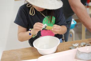 白い帽子の女の子が白いボウルの上で滑石を磨いて勾玉を作っている様子を撮影した写真