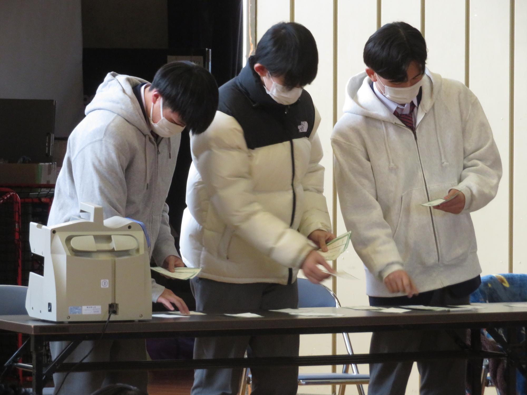 長机の上で投票された用紙を開票作業している3名の男性の写真