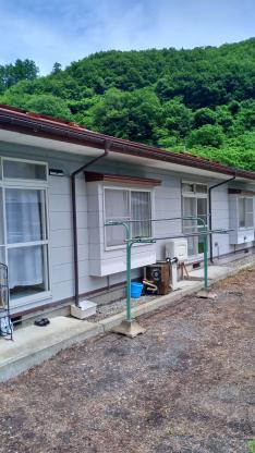 白い外壁と赤い屋根の長屋と物干し台、その奥に見える青々とした木々が生い茂っている山の写真