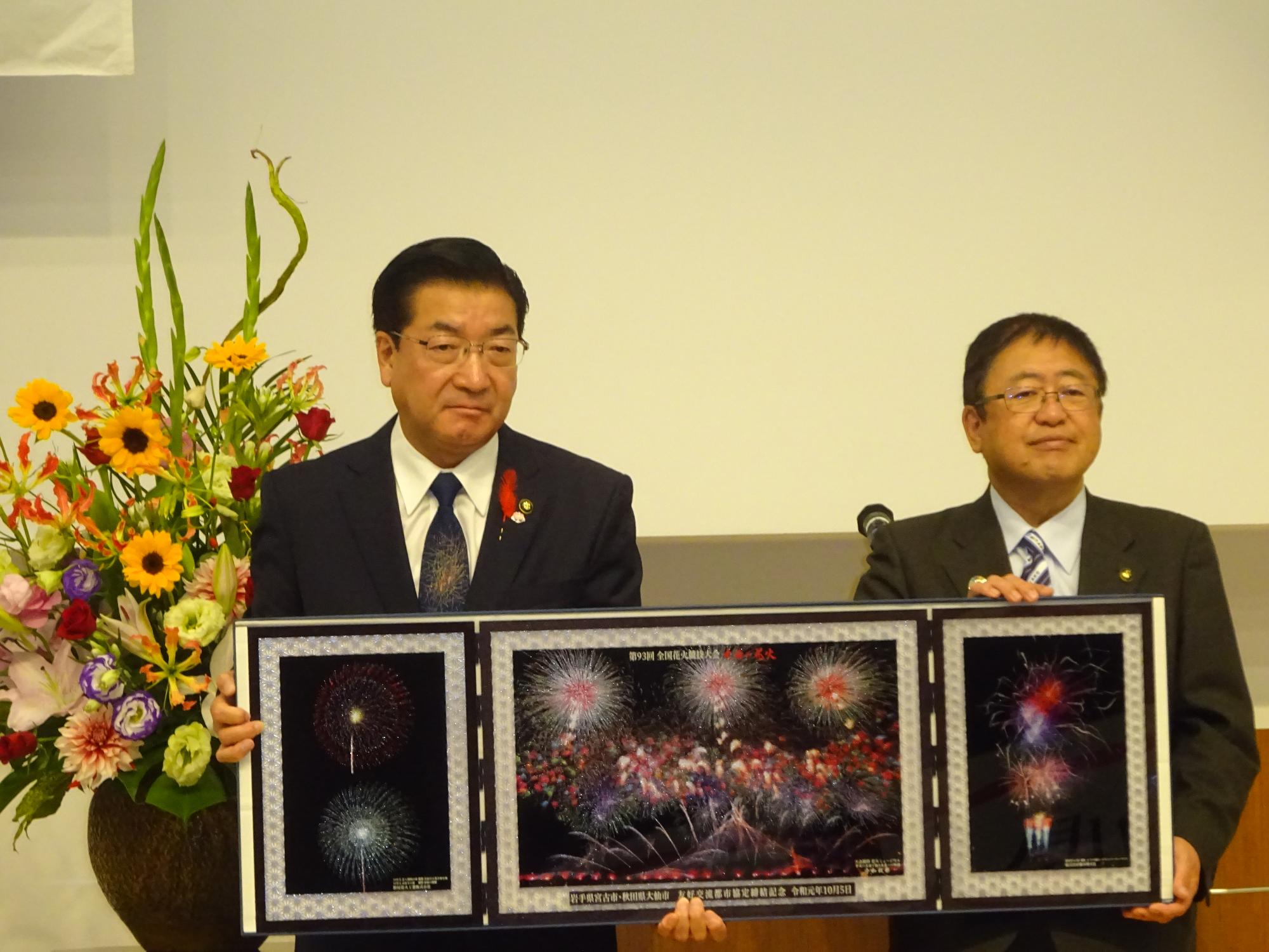 老松大仙市長と山本市長が両手に持つ黒く細長い額縁に、暗闇に色鮮やかな色合いで浮かび上がる花火の写真が飾られている様子の写真