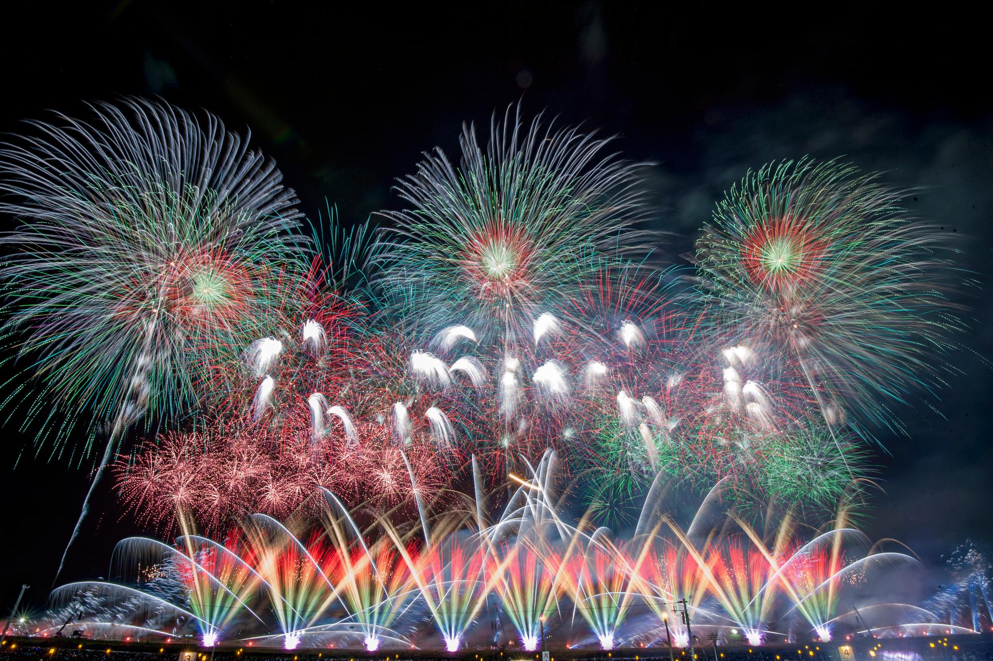 真っ暗な夜空を背景に、大きな花が開いた変化菊やスターマイン、地上から噴出される虹色の鮮やかな創造花火が咲き乱れている写真