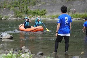 足首が浸かる程の浅瀬で水色のゼッケンをつけた2名がオレンジ色のボートに乗ってオールを漕いでいる様子を2名の関係者が待ち構えているラフティングの写真