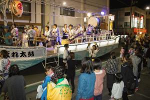 道路中央に船があり、その上で太鼓などの演奏をしているお祭りを沿道で見ている観客たちを写した写真