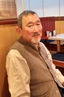 食事をするお店の店内のようなところで、ひげを生やした男性が座ってほほえんでいる写真