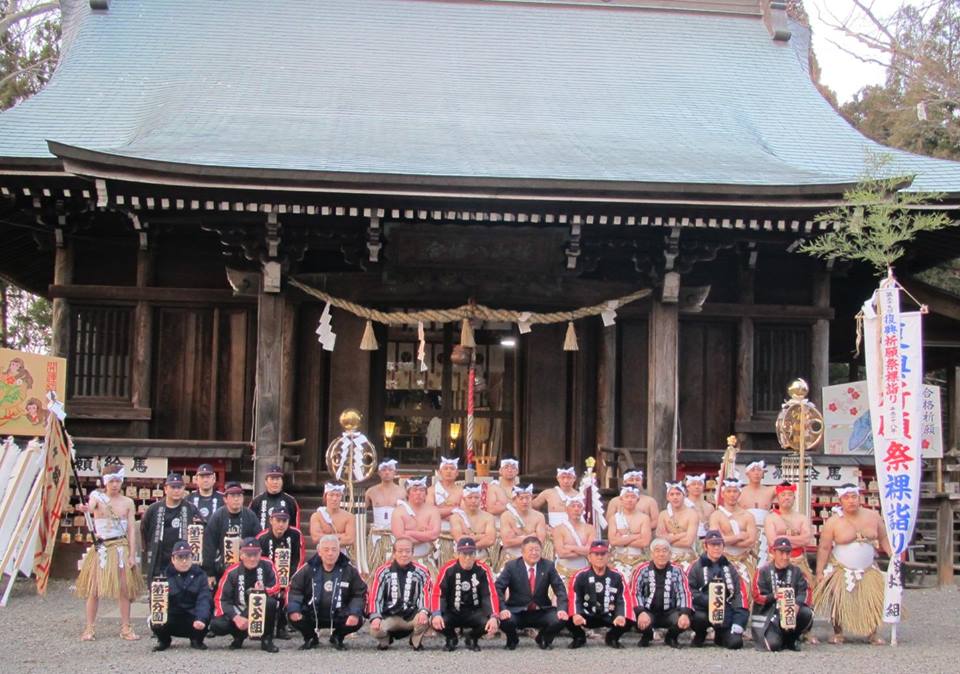 神社の前で上半身裸で鉢巻と腰蓑を巻き両腕を胸の前で組む男性たちと、法被を来た男性たち、スーツを着た市長が3列になって並んでいる記念写真