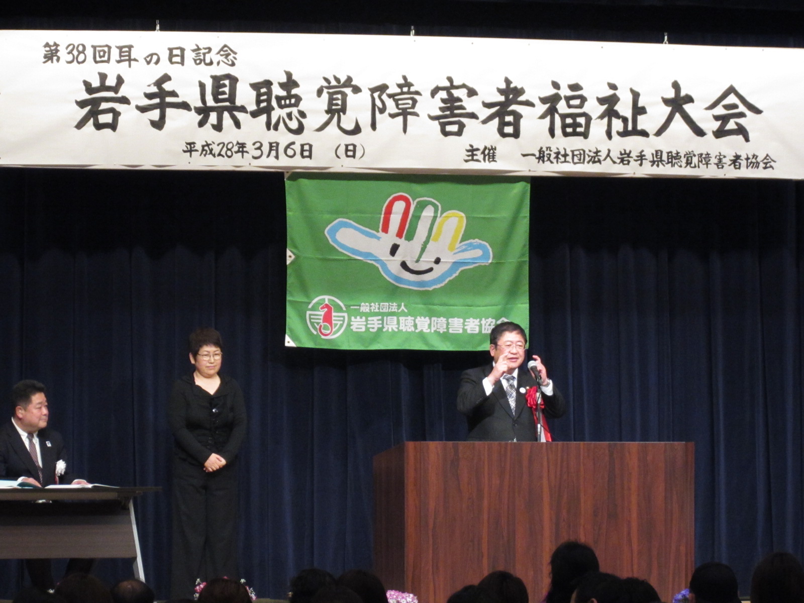 「岩手県聴覚障害者福祉大会」と書かれた横断幕が掲げられた舞台上で、両手を顔の前に出している市長の写真
