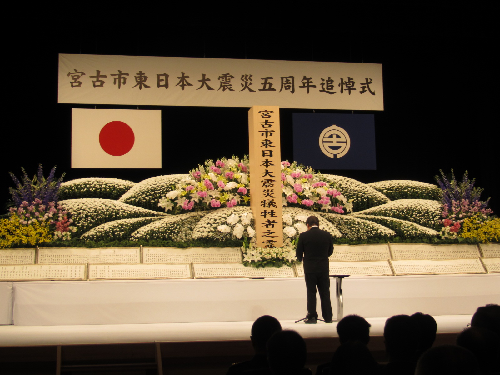 「宮古市東日本大震災五周年追悼式」と書かれた横断幕、宮古市の市町村旗が掲げられ、白色、ピンク色、黄色などの色とりどりの花が飾られた献花台の前で、話をする様子の市長の写真