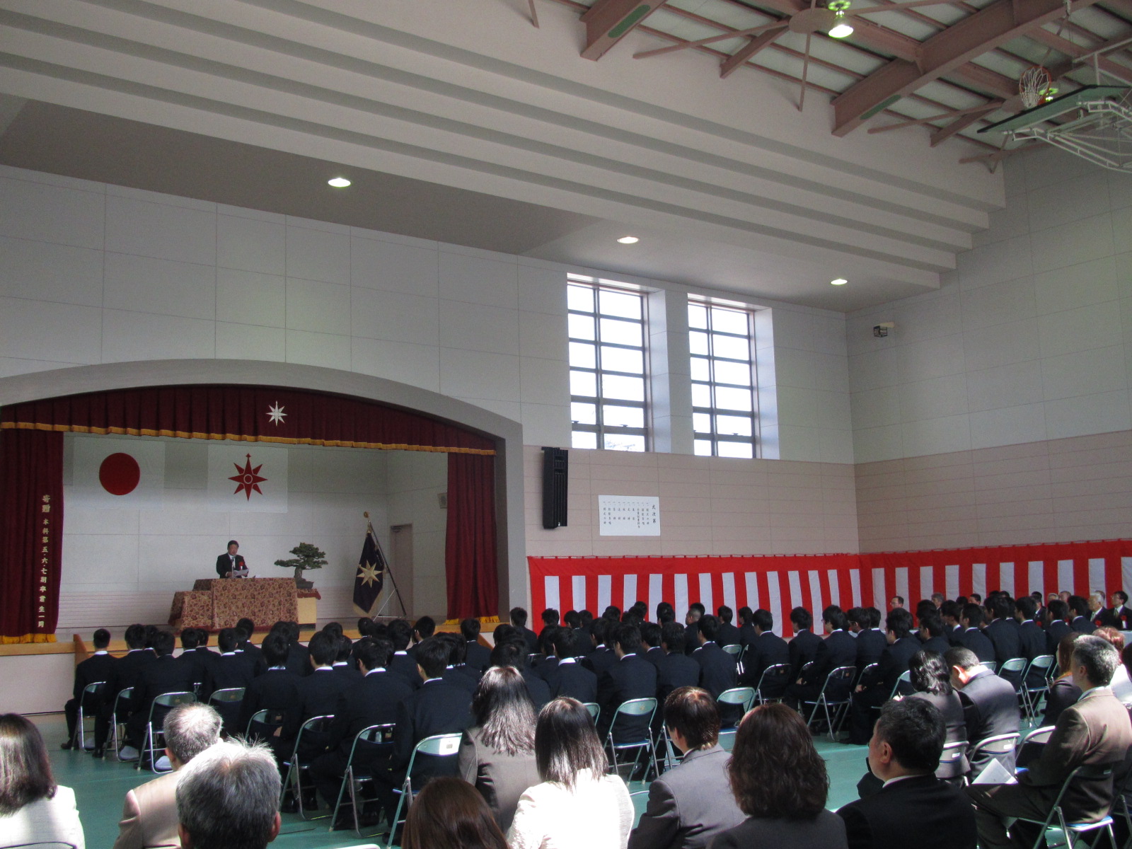 パイプ椅子に座る卒業生たちと、舞台上の演台のマイクに向かい話をする市長の写真