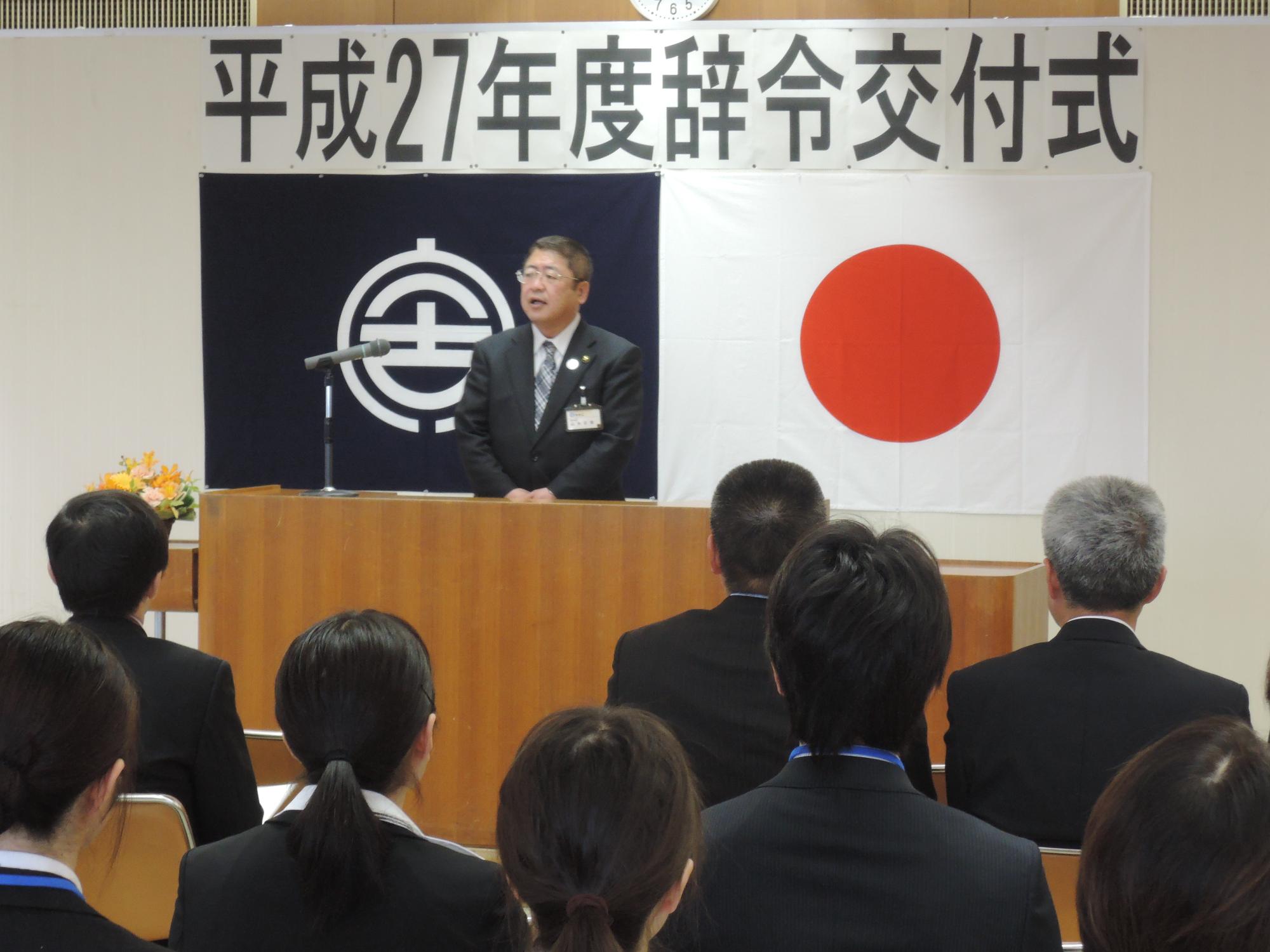 平成27年度辞令交付式と書かれた横断幕の下で、前の演台で式辞を述べている市長と座って聞いて職員の写真