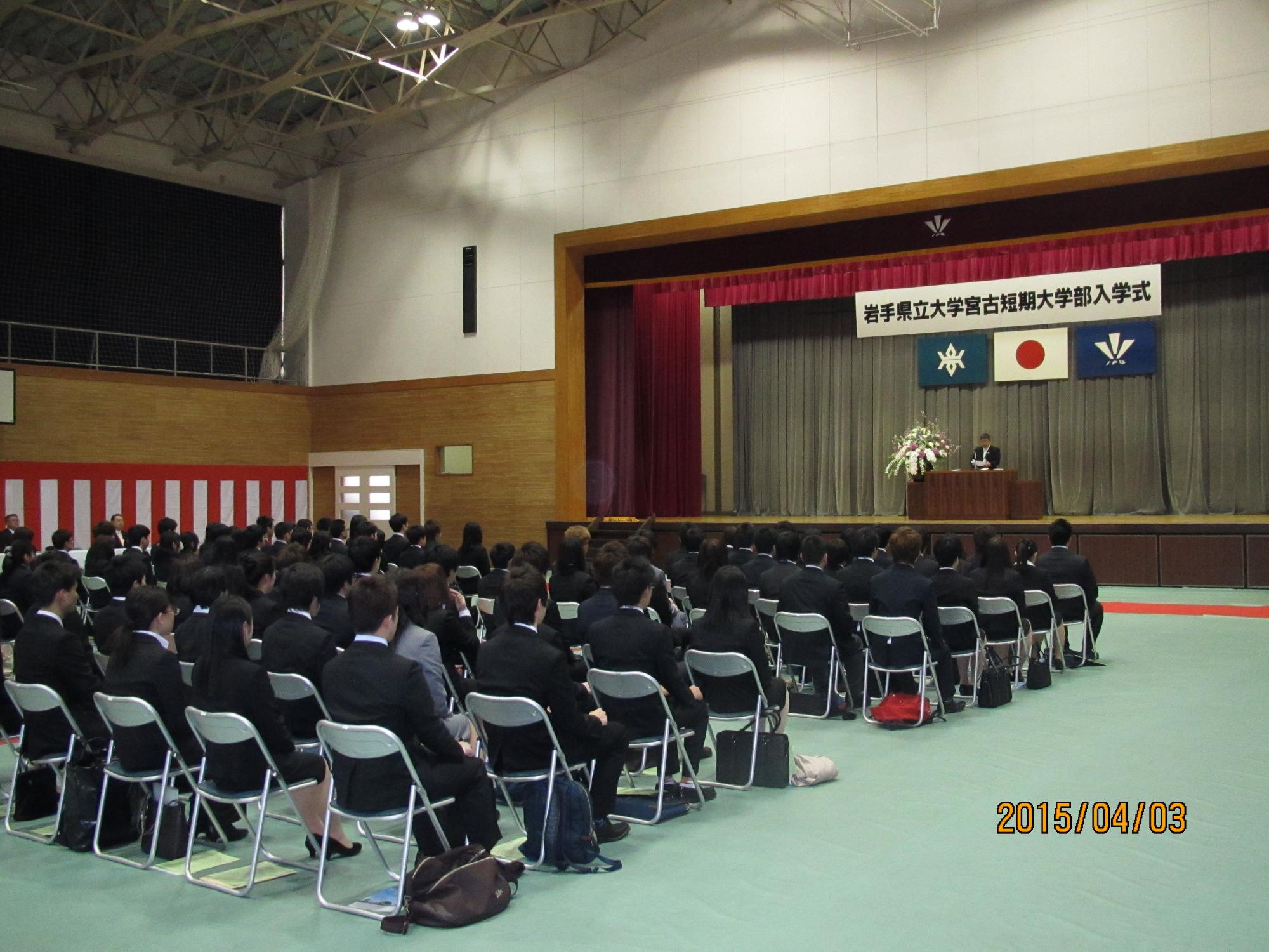 壁に紅白の幕が張られ、岩手県立大学宮古短期大学部入学式と書かれた横断幕のステージ上で、前で祝辞を述べている市長と、座って聞いている入学生たちの写真