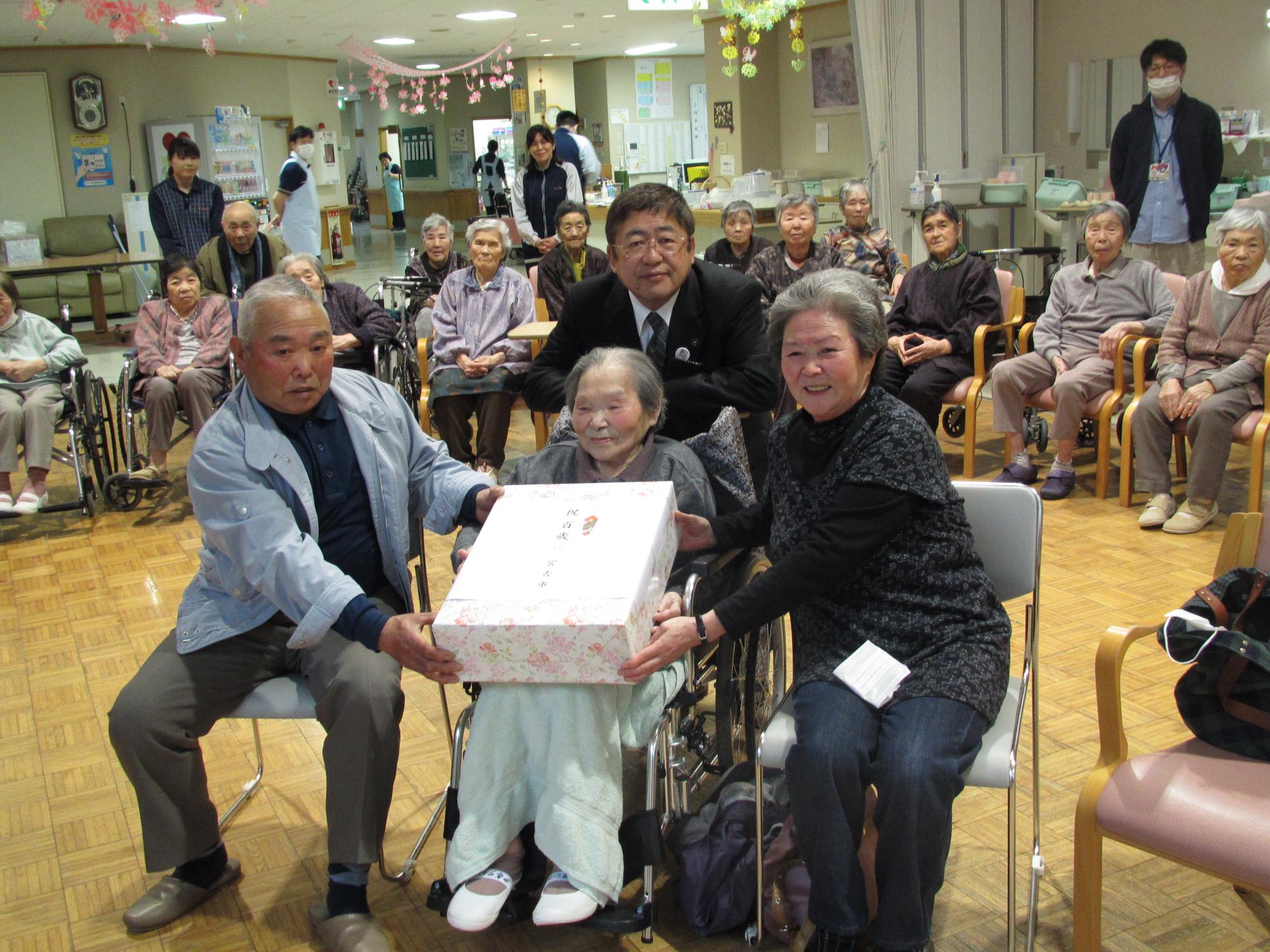 市からの100歳の記念品を久保田シウさんの両脇でご家族が持ち、市長が久保田さんの後ろに立っている光景を施設に入居されている方々、スタッフの方が見守っている写真
