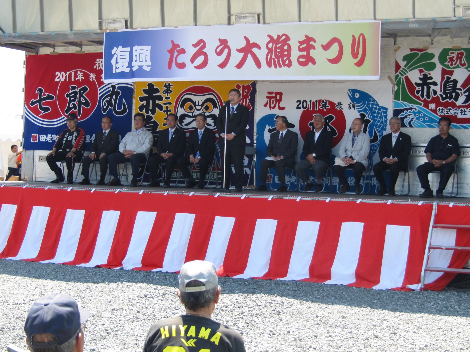 「復興たろう大漁まつり」と書かれた横断幕が掲げられた舞台で、壁に飾られた大漁旗の前で椅子に座る関係者の方々と、スタンドマイクに向かい話をする市長の写真