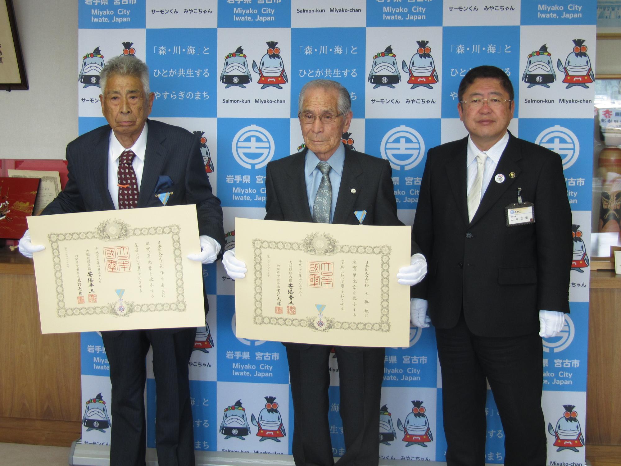 バックパネルの前で賞状を持つ男性2名と市長が並んで立つ記念写真