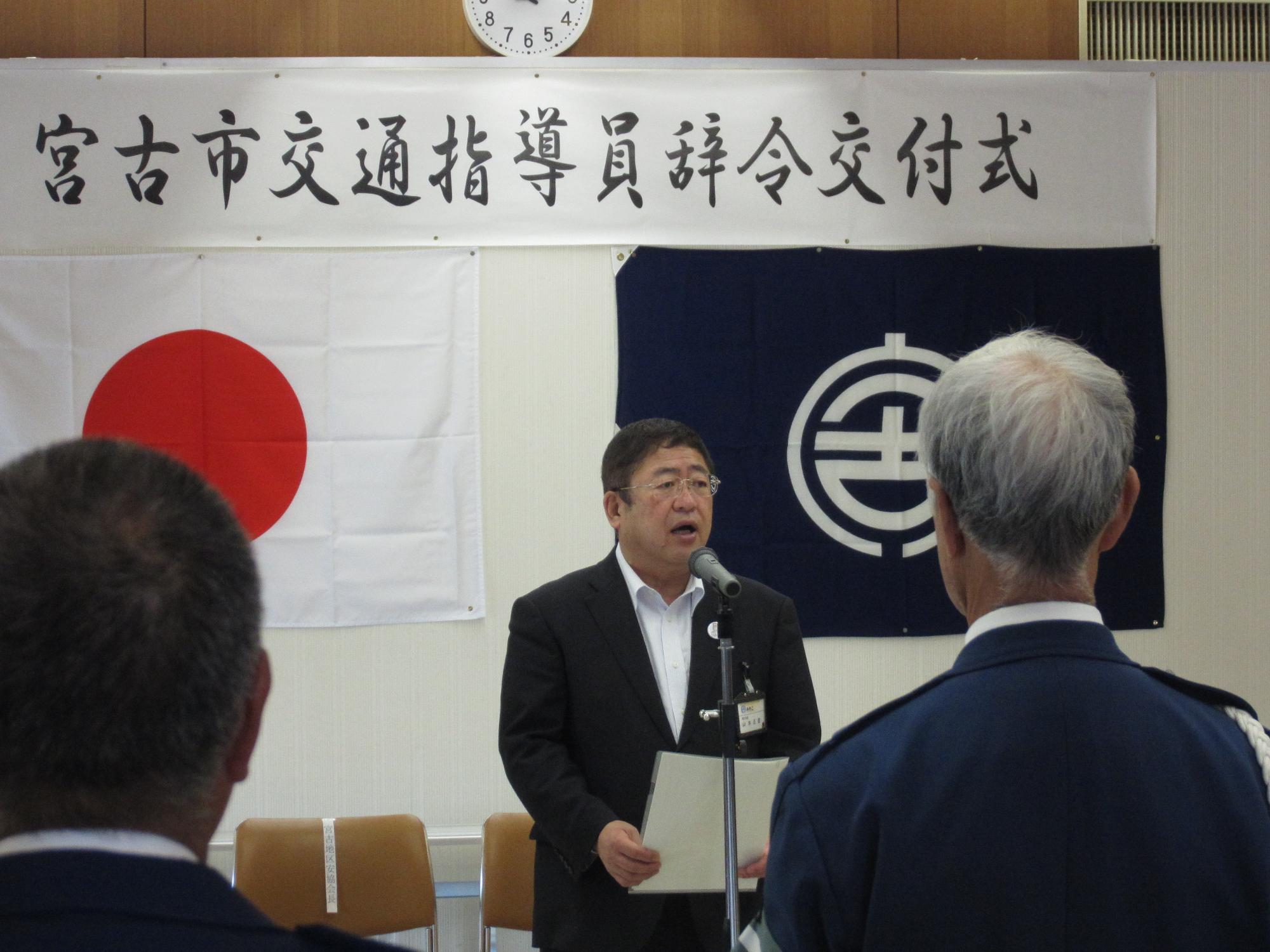 「宮古市交通指導員辞令交付式」と書かれた紙と日本国旗、宮古市の市町村旗が貼られたボードの前で、スタンドマイクに向かい話をする市長の写真