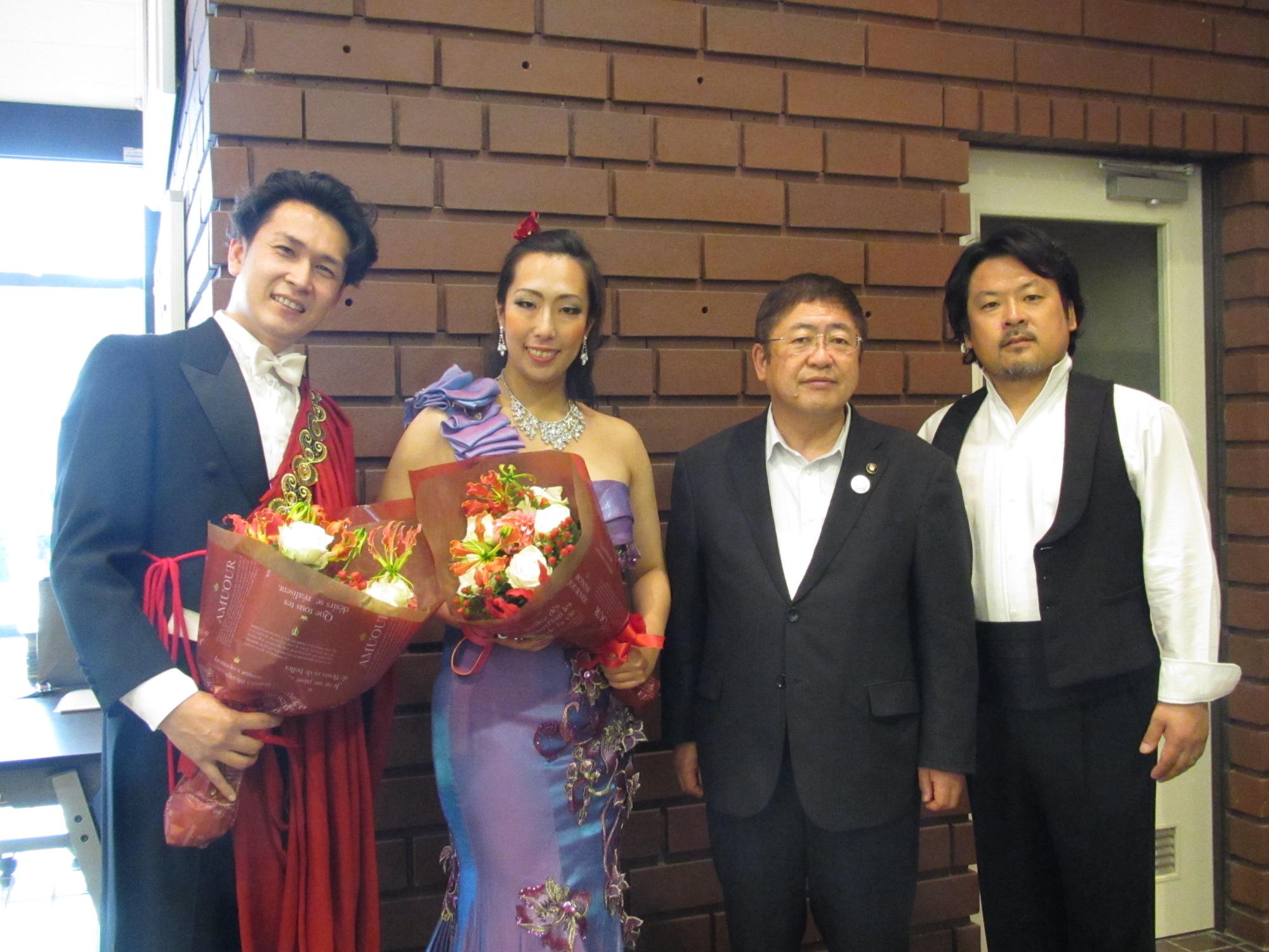 スーツに赤い装飾が施された衣装を着て花束を持つ男性、紫色のドレスを着て花束を持つ女性、スーツを着た市長と黒いベストを着た男性が並んで立つ記念写真