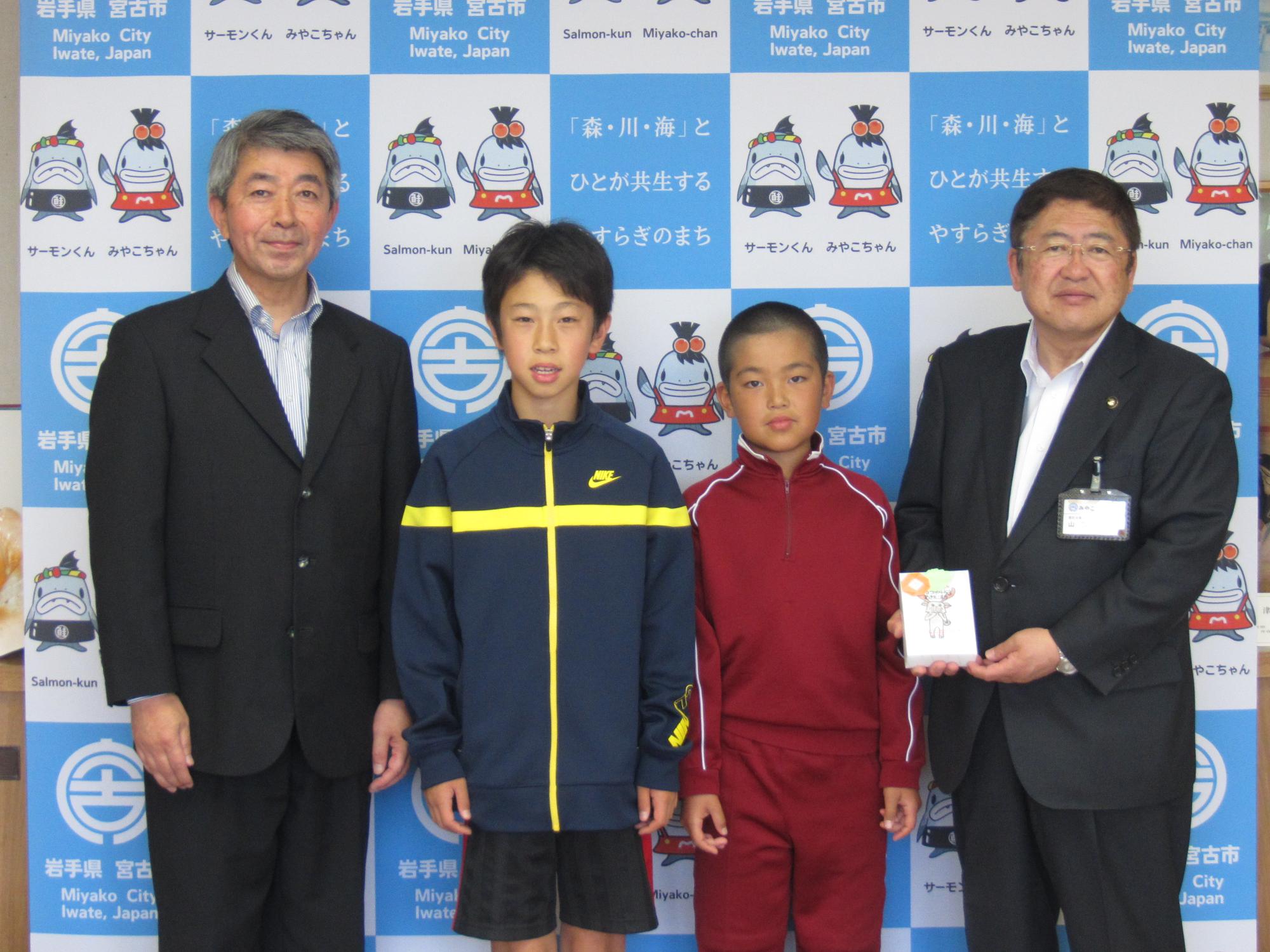 バックパネルの前に立って並ぶ男子小学生2名、関係者の男性1名と小さな箱を持った市長の記念写真
