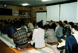部屋の中でたくさんの人が整列して座りながら広聴を聞いている写真